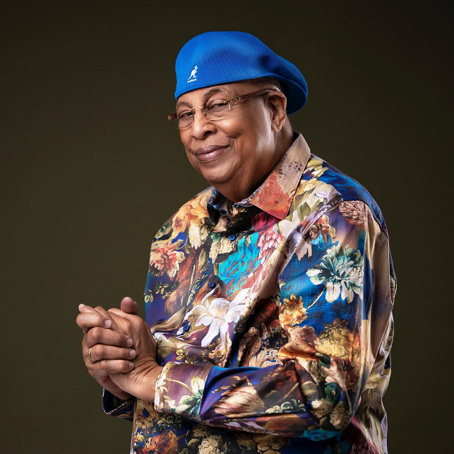Chucho Valdés with folded hands dressed in a floral shirt and blue cap